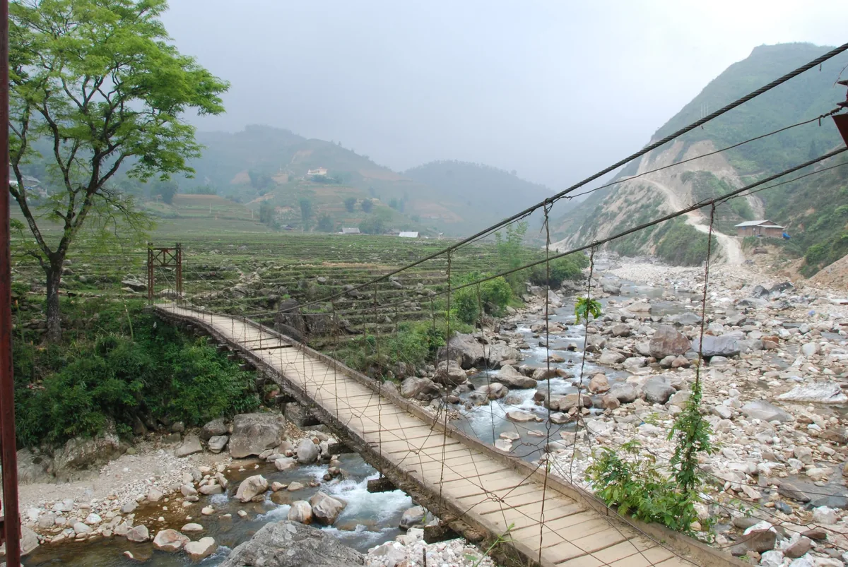 Cầu treo gỗ bắc qua lòng suối đá, phía xa là ruộng bậc thang và núi mờ sương ở Lai Châu
